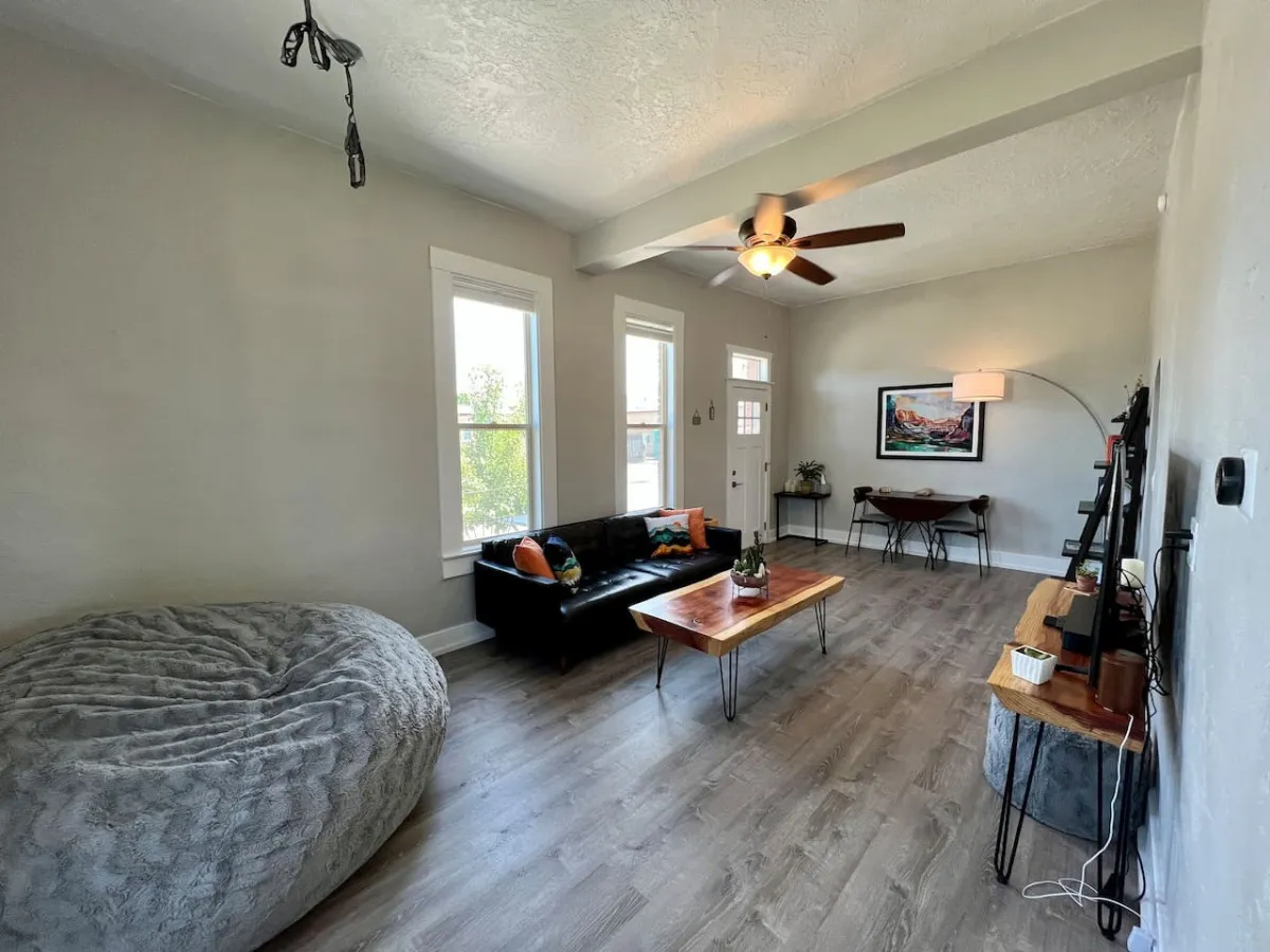 A stylish, modern living room with a black leather sofa, wooden coffee table, bean bag chair, striking wall art, and a ceiling fan. Ample natural light pours in from large windows, creating a serene and welcoming space for relaxation or social gatherings.