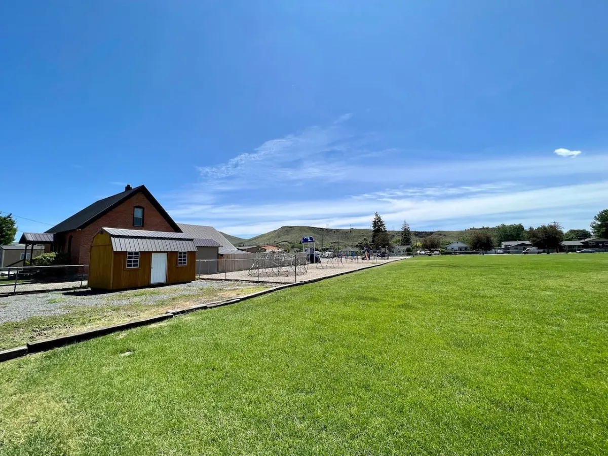 The image displays a green lawn, red brick house, yellow shed, and a playground area, highlighting community, vibrancy, and proximity to a school, with children’s play and family gatherings in a cheerful atmosphere.