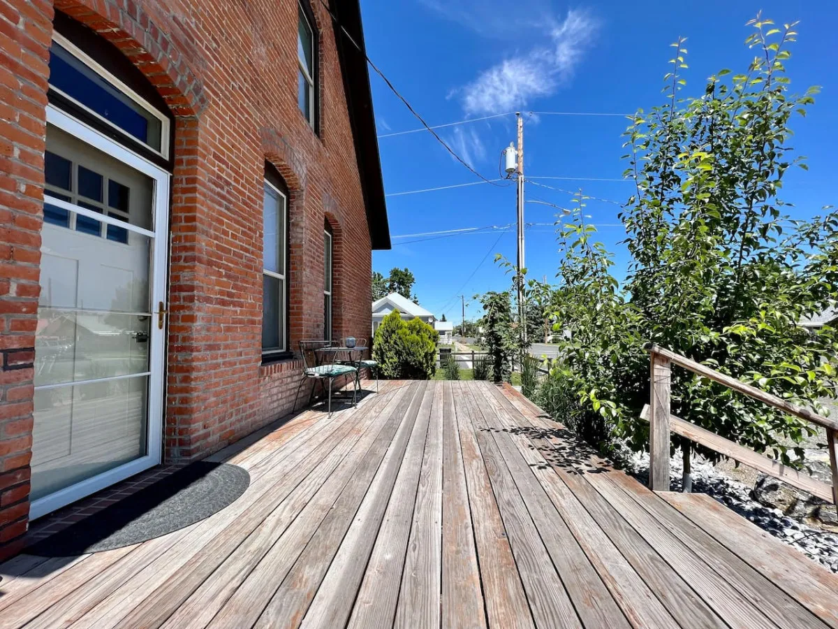 An exterior view of a brick house with a wooden deck and an outdoor seating area, emphasizing the deck as a social space. The setting is bathed in warm natural light, suggesting potential for sunset views or relaxed mornings.