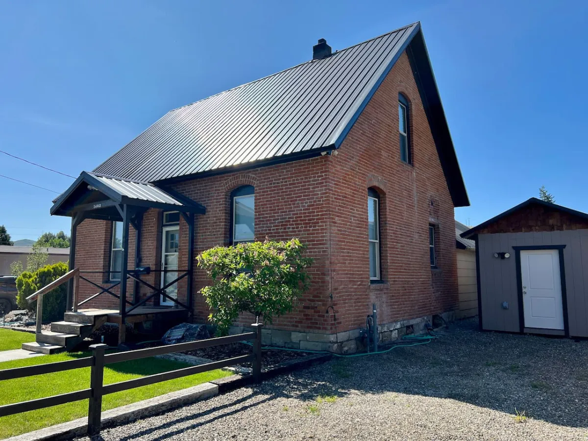 This image showcases the brick exterior of the house, a metal roof, a welcoming front porch with steps, lush green shrubbery, and a wooden fence. The setting evokes a classic and tranquil neighborhood vibe, ideally suited to the section's mention of timeless character and privacy.