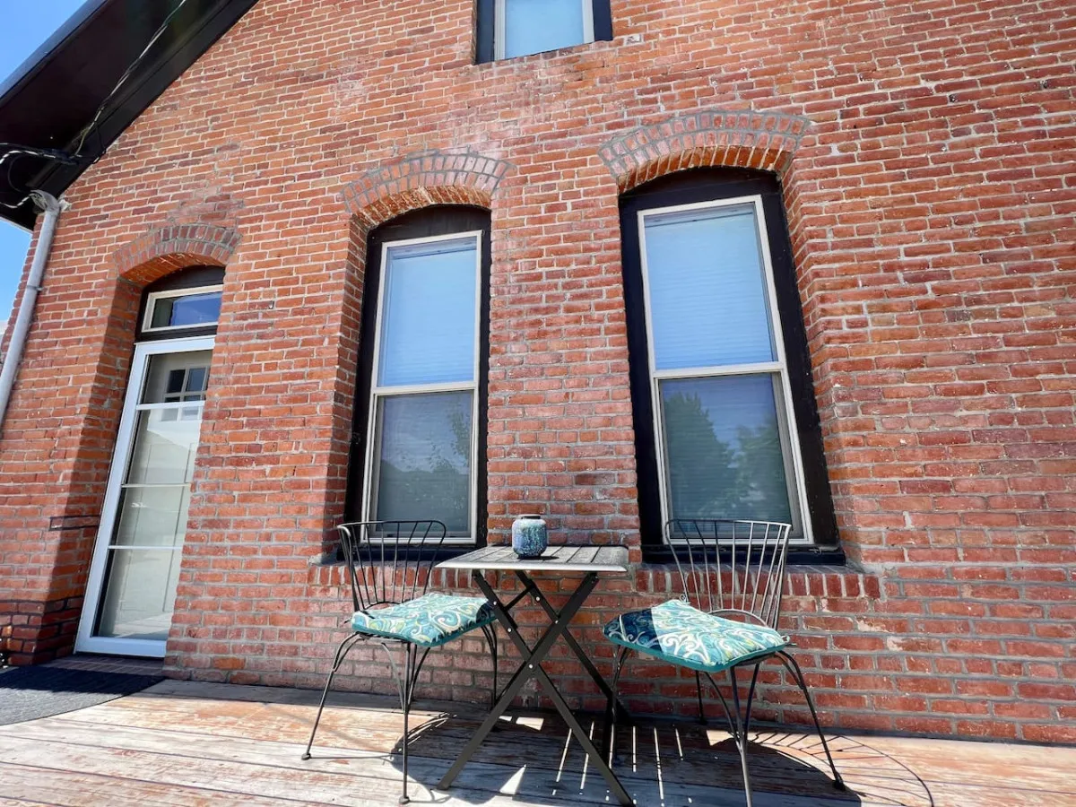 This exterior shot captures a private patio space with a brick wall backdrop, a table and chairs, a decorative cushion, and a planter. The area is cozy and inviting, ideal for outdoor dining, relaxing, or socializing.