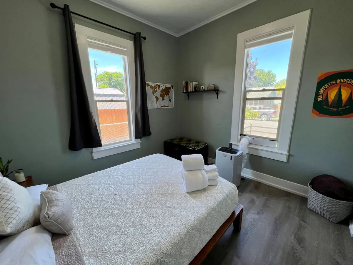 A tranquil queen bedroom with white linens and pillows, a wall-mounted map, shelves with books, and large windows admitting abundant natural light. The setting is both restful and inviting, with simple yet tasteful décor.