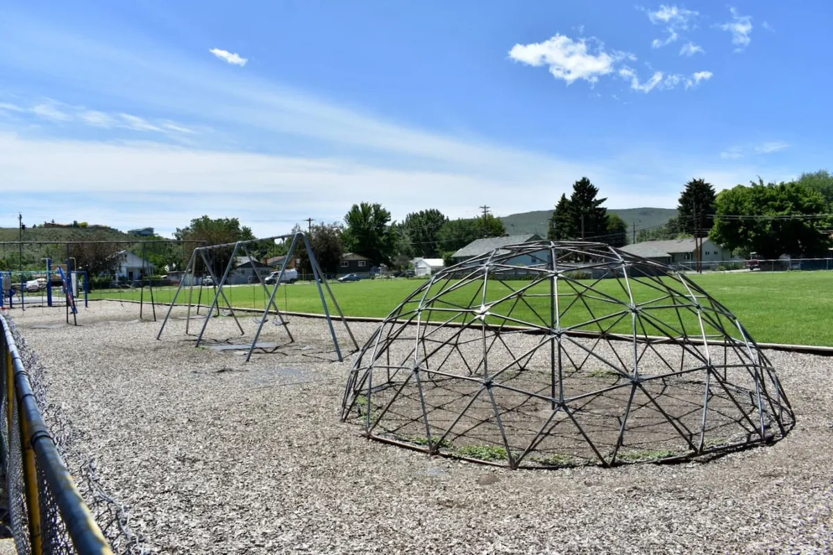 A lively and inviting green field featuring playground swings, a geodesic climbing dome, and ample open space for running and picnics. Children are shown playing and families enjoy the park-like setting, emphasizing communal outdoor fun.