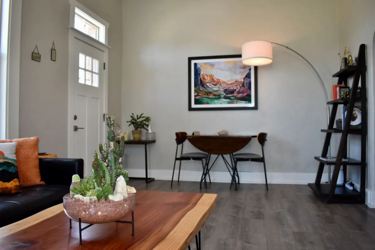 A vibrantly decorated living area with a sofa adorned with colorful cushions, a wooden dining table and chairs, a lush plant arrangement, and large artwork on the wall. The space exudes an inviting, cozy feel ideal for relaxing, dining, or enjoying art.