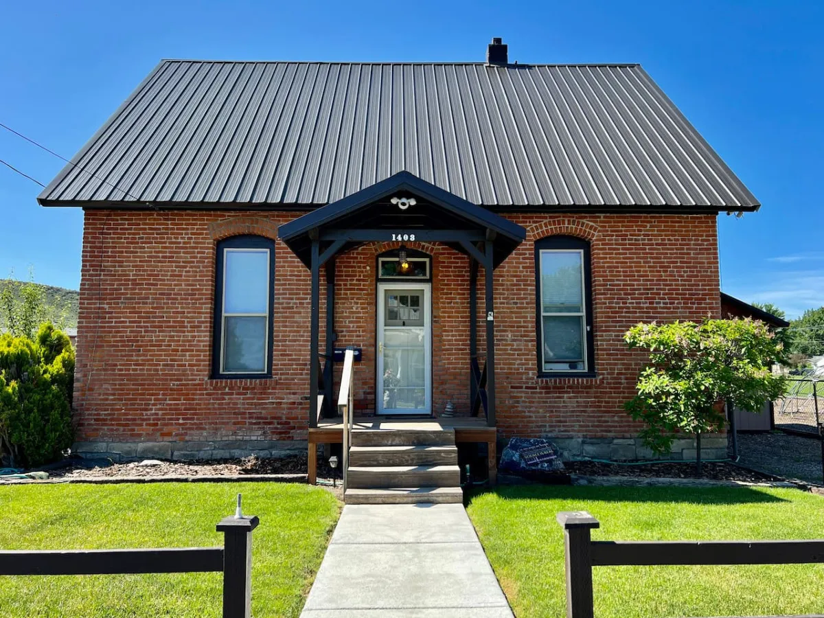 The image showcases the charming exterior of a recently remodeled brick single-family home with a sturdy metal roof. The inviting front porch overlooks a beautifully landscaped yard, all bathed in the warm glow of sunset. The setting evokes a peaceful, welcoming atmosphere ideal for relaxing or enjoying quality time outdoors.