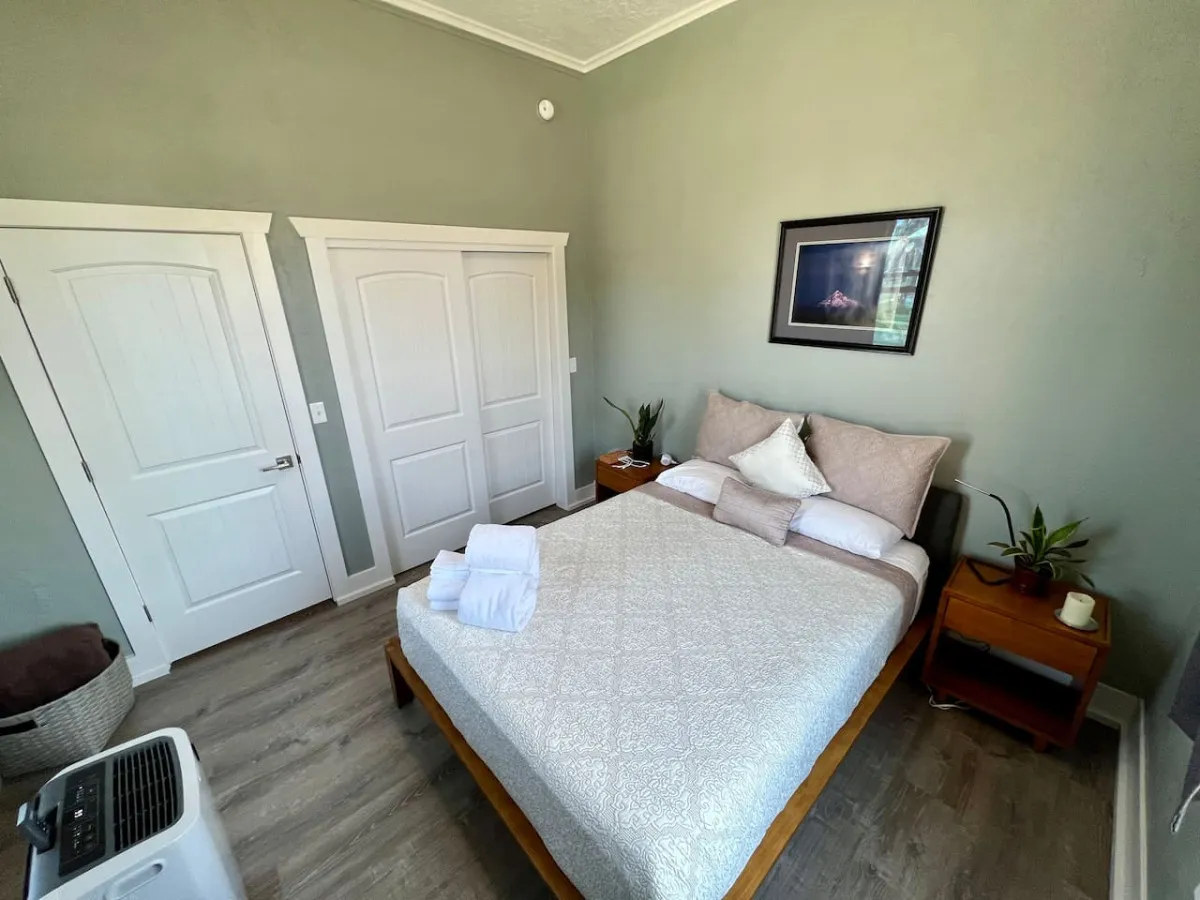 A cozy bedroom scene highlighting a bed with decorative pillows, flanked by two wooden doors and a nightstand adorned with a plant and mug. The muted tones and soft textures evoke comfort and relaxation.