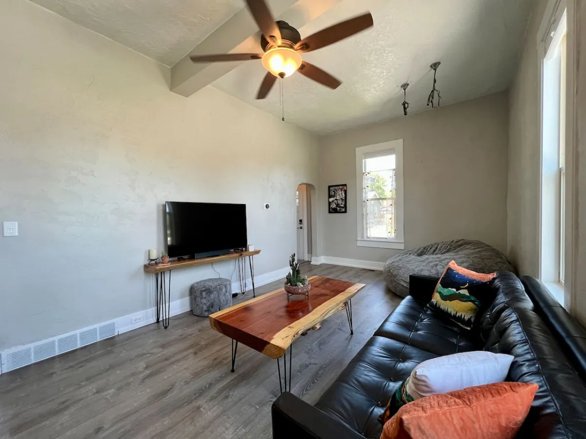 A sunlit living area featuring a black leather sofa, wooden coffee table, television, and a cozy bean bag chair positioned near large windows. Natural light fills the room, creating a comfortable and casual atmosphere ideal for relaxation, reading, or watching TV.
